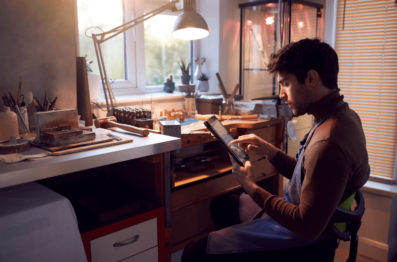 Man sitting in his jewelry workshop, working on a tablet using Gem Logic software to check customer orders. Man sitting in his jewelry workshop, working on a tablet using Gem Logic software to check customer orders.